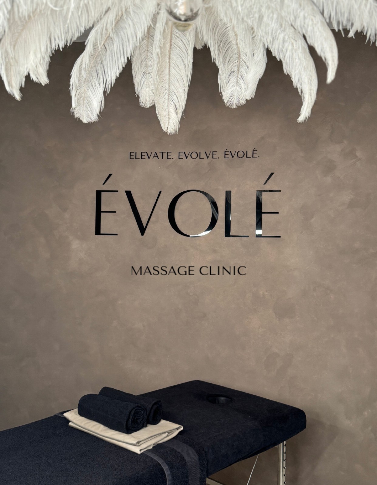 Évolé Massage Clinic treatment room with feather chandelier, ÉVOLÉ branding, and massage table